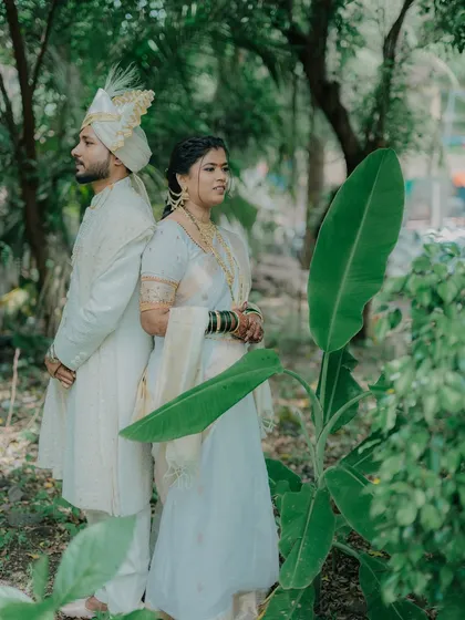 A serene portrait of a couple in matching white traditional outfits, standing back-to-back in a quiet, green grove. This pose creates a sense of partnership and shared strength, highlighting their elegant Maharashtrian wedding look.