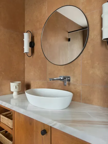 In the East Coast Residence, this bathroom showcases our focus on integrating storage without compromising on style. The wooden vanity with open shelving provides functionality, while the warm-toned tiles and unique sconces add character.