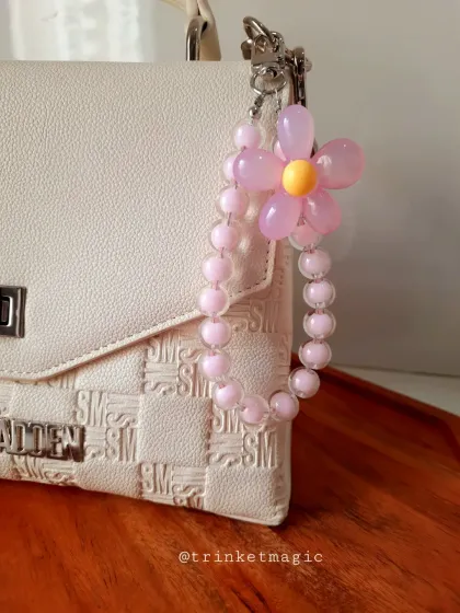 This handmade pastel flower charm adds a delicate, feminine touch to a white handbag. The pink beaded loop and flower make it a perfect accessory for a soft, aesthetic style.
