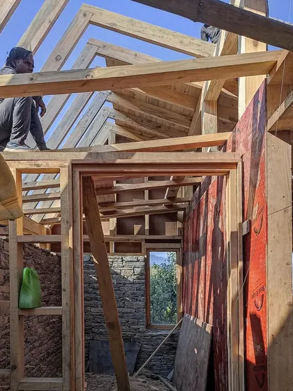 An interior view during the framing stage. A carpenter sits atop the wooden roof structure, highlighting the hands-on nature of the build and the intricate joinery required for mountain architecture.