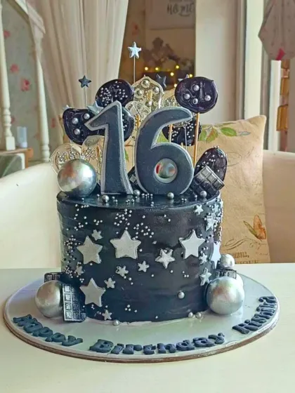 A sleek and stylish black cake for a 16th birthday, decorated with silver stars, chocolate bars, and metallic spheres.