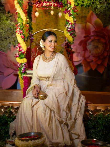 Actress Kavya Gowda seated at a beautifully decorated event, her elegant white Anarkali standing out against the floral backdrop.