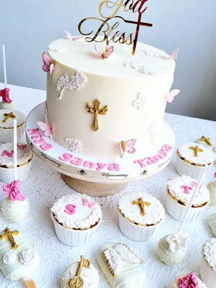 A Holy Communion cake with matching cupcakes and cakesicles. The design is elegant and simple, with white buttercream, gold crosses, and delicate pink butterflies.