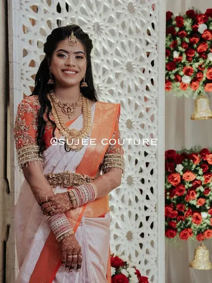 The pure joy of a bride on her wedding day. She is wearing a custom orange and white Kanchipuram saree, complemented by a hand-embroidered blouse with detailed work on the sleeves, designed to make her feel special.