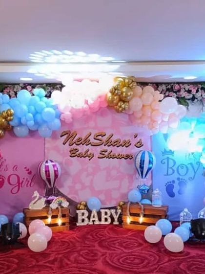 A classic 'It's a Girl / It's a Boy' gender reveal baby shower stage. The decoration uses pink and blue balloons, hot air balloon motifs, and 'BABY' blocks to build anticipation.