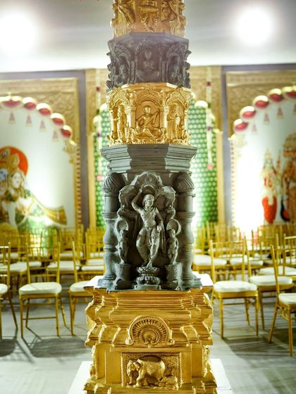 A close-up of a custom-designed pillar, showcasing the detailed carving work inspired by South Indian temple architecture. The combination of faux grey stone and gilded gold elements adds a sense of authenticity and grandeur.