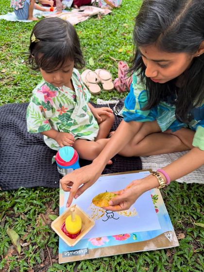 A mother and daughter duo work together on a nature painting. The mother guides her child's hand, showing her how to use pigments made from flowers to create art.
