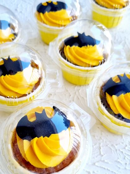 Batman-themed cupcakes with yellow buttercream and a fondant bat symbol, presented in individual containers.