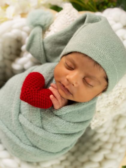 A close-up shot of a sleeping baby in a sage green wrap, holding a small red heart. This simple pose highlights the baby's peaceful features.