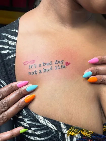 A closer look at the fine-line quote tattoo "it's a bad day, not a bad life" done in red and black ink, perfectly complemented by the client's bright, summery nails.