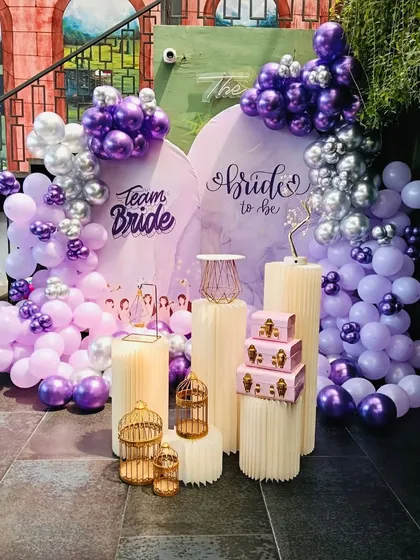 I had so much fun creating this 'Team Bride' setup for a bridal shower. The decor features a custom backdrop with a beautiful balloon arrangement in shades of purple, lilac, and silver. The different-sized pedestals and gold birdcages add a touch of whimsy and are perfect for placing gifts or desserts.