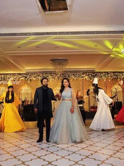 The couple is welcomed by a troupe of colorful Sufi dancers. The vibrant costumes against the elegant white and gold decor of the ballroom create a stunning visual contrast.