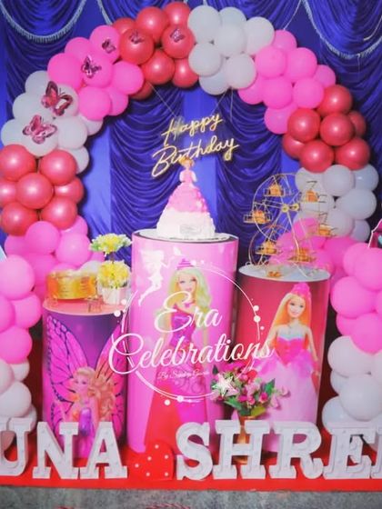 A pink Barbie-themed first birthday party. The decoration includes character-printed plinths, a pink balloon arch, and a light-up number '1' for a dreamy celebration.
