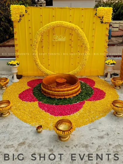We created a stunning floor-based Haldi setup for this rooftop event in Kalaburagi. The decor features a large floral rangoli made from yellow, pink, and green petals, with a traditional brass "urli" at its center. The backdrop is a simple yellow drape with a marigold wreath, keeping the focus on the intricate floor work.