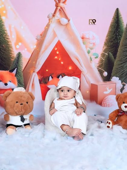 A baby rests in a beanbag chair, surrounded by teddy bears, a unicorn, and a fox in a snowy woodland camp. The warm glow from the teepee creates a cozy and magical winter atmosphere.