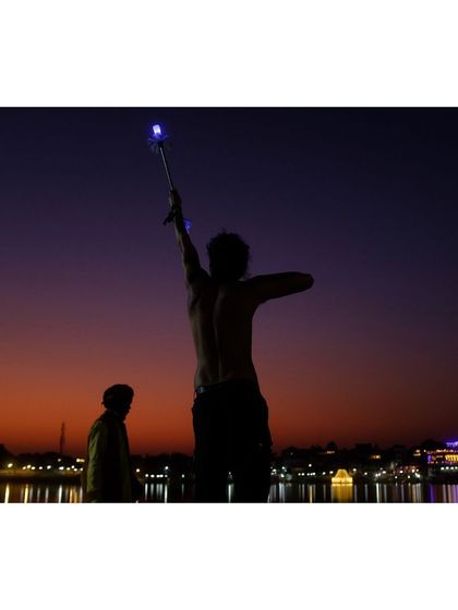 The juggler raises a glowing stick to the sky in a triumphant pose. This silhouette shot captures the dramatic conclusion of his performance by the lake.