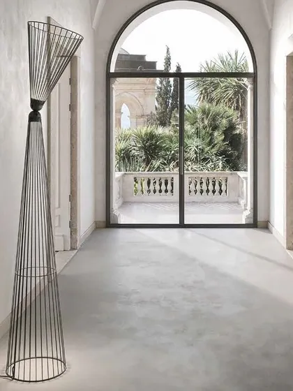 The tall, elegant form of the Ikonika floor lamp in a covered hallway, looking out onto a balcony. This piece is part of the "Collection of Crossed Destinies" and is suitable for both indoor and outdoor use.