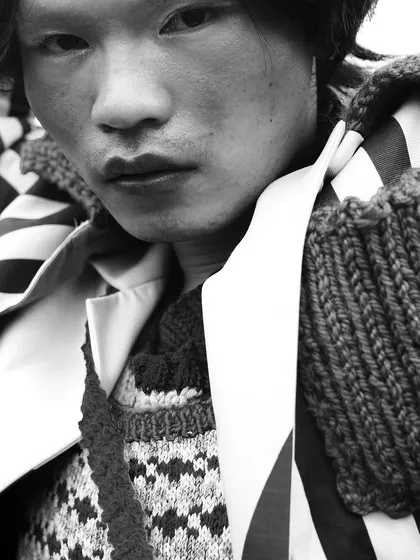 A powerful black and white portrait from the "RIOT" series. The tight crop focuses on the model's expression and the intricate textures of the layered knitwear and striped collar.