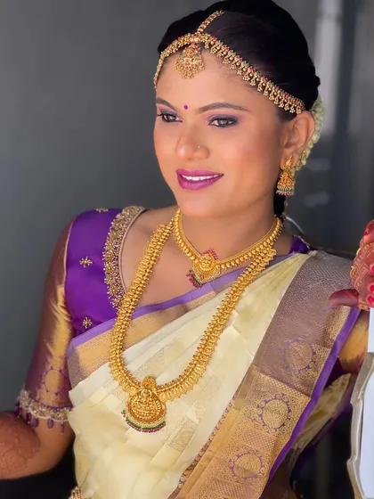 A beautiful bride in a classic off-white and purple Kanchipuram saree. Her makeup features a striking purple eyeshadow to match her blouse and a radiant, glowing complexion.
