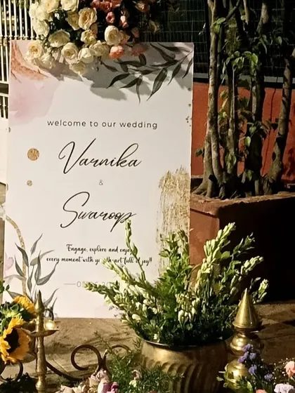 A personalized welcome sign for a wedding, adorned with delicate watercolor-style florals. The sign is placed on an easel and surrounded by brass pots and fresh flowers, adding a touch of elegance to the venue entrance.