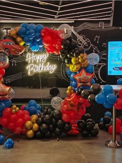 A high-energy Disney Cars themed birthday party. The decoration includes a circular balloon arch in red, black, and blue, Lightning McQueen foil balloons, and a central 'Happy Birthday' neon sign.