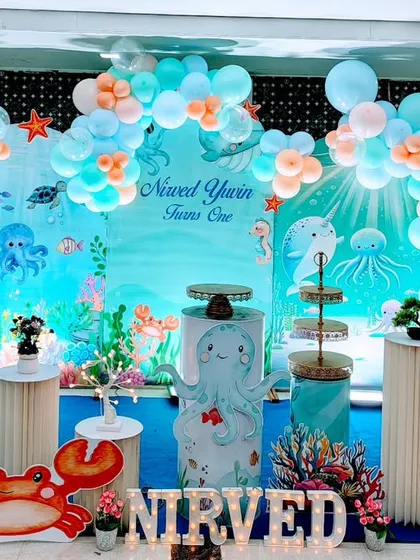 An immersive under the sea aqua theme for a first birthday celebration. This vibrant setup features a custom ocean backdrop, themed cake stands, character cutouts of a crab and turtle, and a balloon garland in seafoam green and pastel blue.