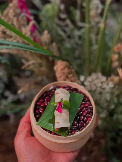 A small wrap, possibly a Peking duck roll, presented in a mini bamboo steamer on a bed of red beans.