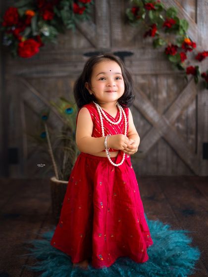 A sweet little Radha in a simple red dress, looking shyly at the camera.