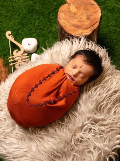 A top-down view of a baby wrapped in a deep orange swaddle, lying on a soft fur rug. The surrounding props like a miniature charpai and a basket of vegetables add to the rustic, village-life theme.