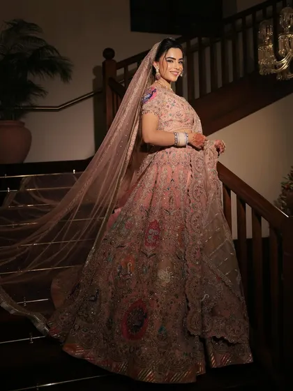 The bride on a grand staircase, her pastel lehenga and long veil creating a dramatic and beautiful silhouette. Her makeup is soft and glowing, fitting for a princess.