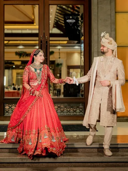 A classic couple portrait on the wedding day, showcasing the bride's stunning red lehenga and the groom's elegant sherwani. This is a timeless shot that captures their regal presence.