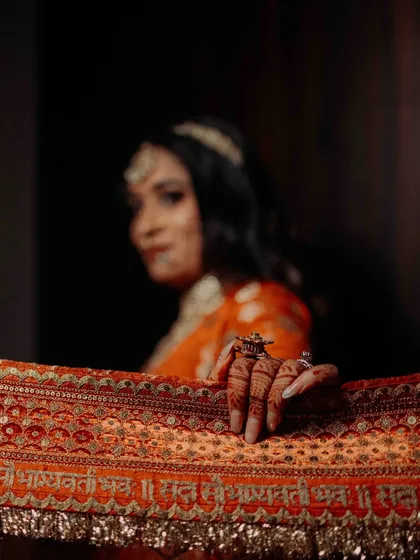 An artistic shot focusing on the bride's hand, adorned with henna, resting on her ornate dupatta. The inscription 'Sada Saubhagyavati Bhava' is visible, adding a traditional blessing.