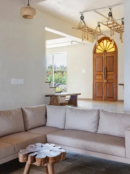The living room features a mix of custom and sourced furniture, including a live-edge wood coffee table and a unique light fixture made from a clothes hanger. The arched wooden door adds a touch of traditional Indian architecture.