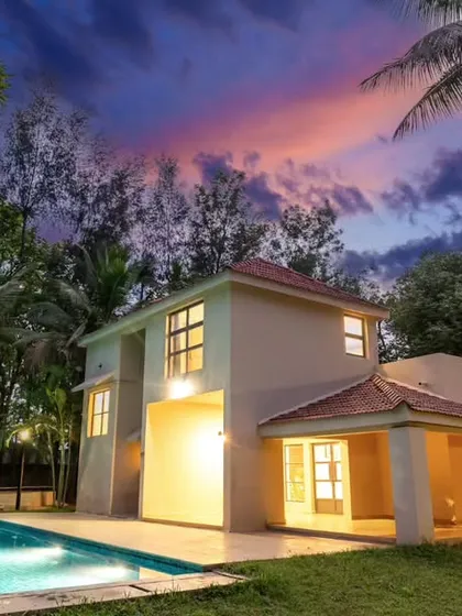 The villa and pool captured from a different angle, with a tall palm tree framing the shot. The warm glow from the house reflects on the water, creating a peaceful and inviting evening scene.