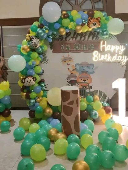A fun jungle-themed first birthday with a circular backdrop and a large light-up '1'. The organic balloon arch in jungle colors and scattered floor balloons complete the wild look.