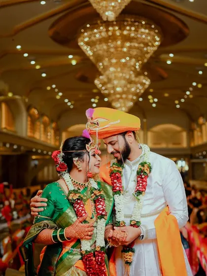 Capturing the grandeur of a traditional Maharashtrian wedding. The couple shares a look under a magnificent chandelier, surrounded by the warmth and opulence of their celebration, a perfect blend of tradition and splendor.
