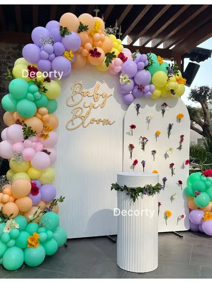 A beautiful 'Baby in Bloom' themed baby shower backdrop. The setup features a colorful pastel balloon arch and a white panel decorated with real flowers for a fresh, floral celebration.