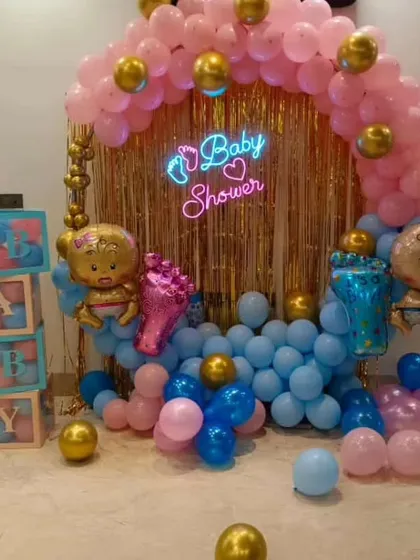 A vibrant baby shower backdrop with a gold tinsel curtain and a 'Baby Shower' neon sign. The setup is framed by a colorful balloon arrangement in pink and blue with gold accents.