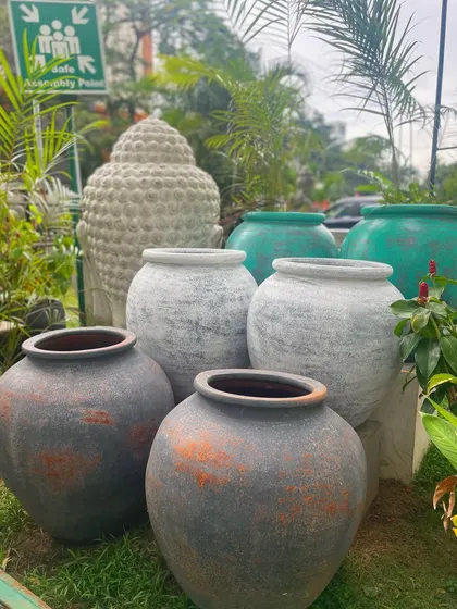 A beautiful assortment of our terracotta and ceramic pots. We offer a range of sizes and finishes, from rustic greys and earthy browns to vibrant sea greens, with a Buddha head statue in the background.
