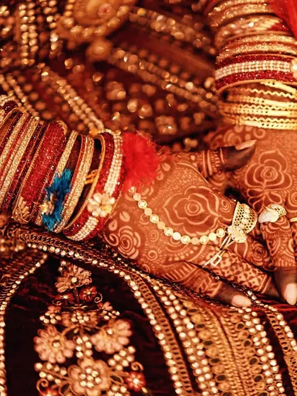 A close-up of a bride's hands, showing the deep maroon stain of the mehndi against her ornate dress and jewelry.