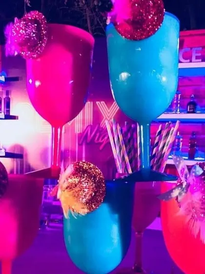 Oversized, brightly colored goblets ready for some disco-era cocktails. The pink and blue glasses, garnished with glittery disco balls, stand out against the neon-lit bar backdrop.