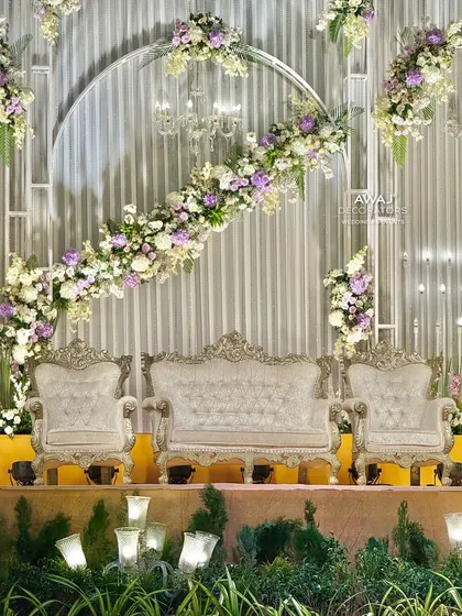 A modern and sophisticated stage design featuring a sweeping arc of white and lavender florals over ornate silver thrones, set against a contemporary silver-paneled backdrop.
