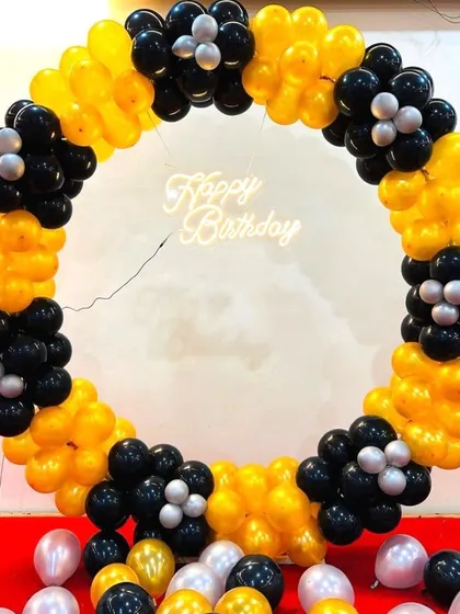 A striking balloon ring decoration in a yellow and black color scheme. The design features flower-like clusters of black balloons and a 'Happy Birthday' neon sign, perfect for a modern party.