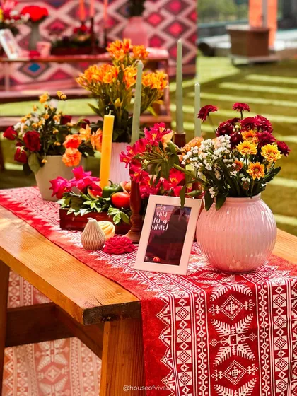 A rustic yet vibrant tablescape featuring a traditional patterned runner, colorful floral arrangements in a pink vase, and tall candles, perfect for a lively daytime event.