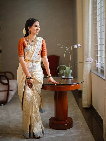 A beautiful, naturally lit portrait of a bride in a classic off-white and rust-orange silk saree. The makeup is kept soft and natural to enhance her features, creating a look of pure elegance.