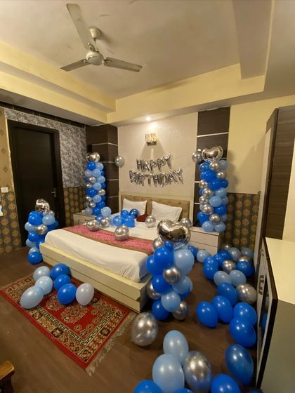 A birthday room decoration in shades of blue and silver. The setup includes balloon pillars by the bed, loose balloons on the floor, and a 'Happy Birthday' foil banner.