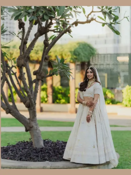 A beautiful full-length portrait of the bride in her elegant white lehenga, standing by a tree in a garden. The soft, natural light and serene setting create a timeless and graceful image.