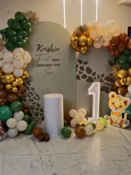 For a child's first birthday, I can create a memorable "First Trip Around the Sun" jungle theme. This setup includes a custom-printed backdrop with a giraffe pattern, surrounded by an organic balloon garland in earthy tones of green, brown, and gold. I also provide character cutouts like a baby giraffe and lion, a light-up marquee number '1', and a pedestal for the cake.