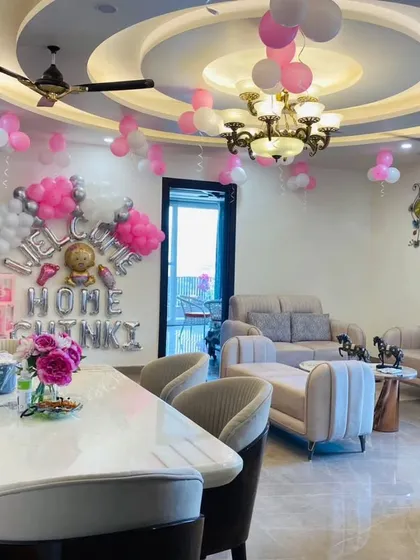 Welcome your little one home to a room filled with love. This is a beautiful living room decoration with "Welcome Home" foil balloons, pink and white balloon clusters on the ceiling, and transparent 'BABY' boxes for a modern touch.