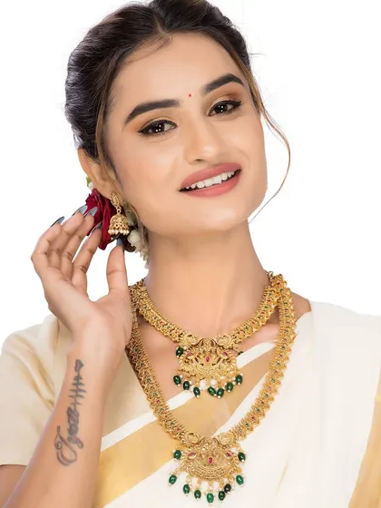 A smiling model presents a traditional gold necklace set with green bead accents. This friendly and approachable shot is perfect for social media content and online stores like Myntra or Ajio.